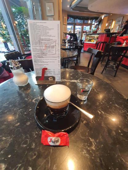 A coffee drink with a thick foam layer sits on a table, accompanied by a small glass of water and a sugar packet. A menu is visible in the background.