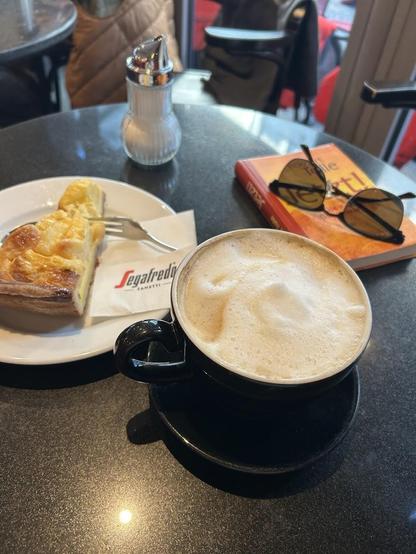 A table setting with a coffee cup, saucer, pastry, sugar dispenser, book, and sunglasses.
