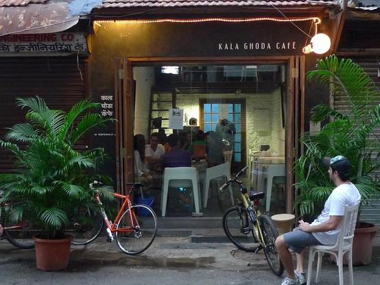 An inviting Kala Ghoda Cafe entrance with people inside. A man sits outside by plants and bicycles.