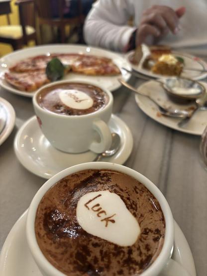 Two cups of cocoa with 'Luck' and 'Good' written in white on the surface. Pizza and pastries are also visible.