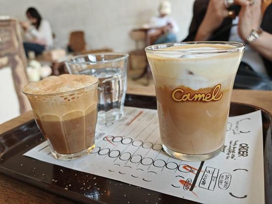 Two coffee drinks and a glass of water on a tray. The taller drink has a "Camel" logo.