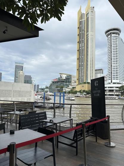 Outdoor seating area by a river with skyscrapers in the background. "The Coffee Club" sign is visible.