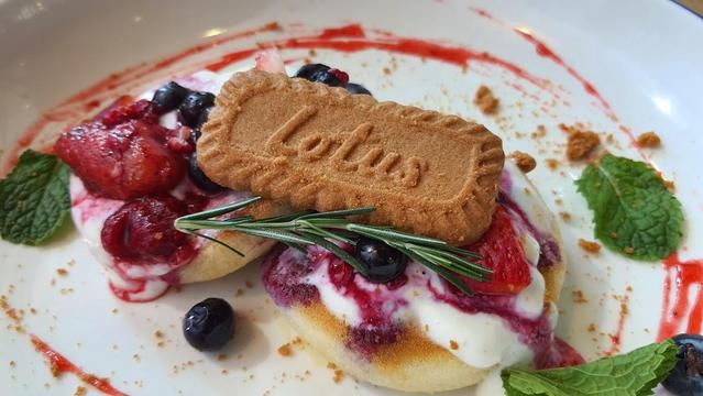 A close-up of a dessert plate with pancakes, berries, cream, a Lotus biscuit, and mint garnish.