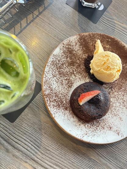 A top-down shot shows a plate with a chocolate lava cake topped with a strawberry slice, a scoop of vanilla ice cream, and a light dusting of cocoa powder. A green drink with ice is to the left of the plate.