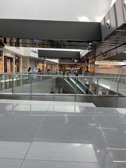 A bright, tiled mall interior with Zara and Pennyblack stores visible. An escalator is present.