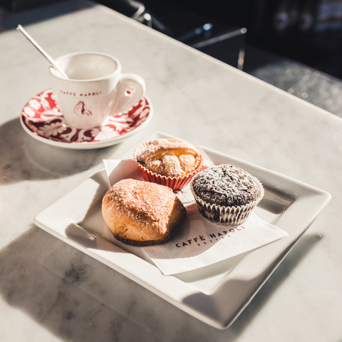 A white plate with three pastries sits next to a Caffe Napoli cup and saucer on a marble surface.