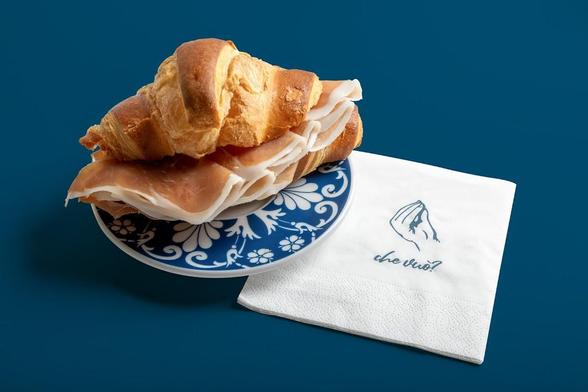A croissant sandwich filled with slices of ham, served on a blue and white patterned plate with a napkin that reads "che vuoi?" on a dark blue background.