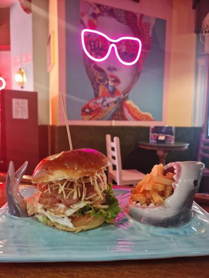 A burger with fries served on a blue plate with a shark motif, against a restaurant backdrop.
