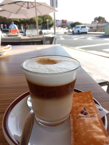 A latte in a glass with a spoon and sugar packet on a saucer.
