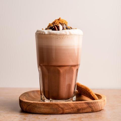 Chocolate drink in a glass topped with whipped cream, chocolate sauce, and cookie crumbs, served on a wooden tray with a cookie.