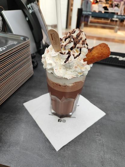A decadent chocolate drink with whipped cream, chocolate drizzle, a cookie, and a wooden stirrer on a napkin.