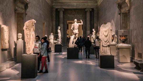 Visitors are in a Greek and Roman gallery at The Met that is filled with large white statues of figures. It's evening and the room is dark with spotlights on the art. Visitors are in a Greek and Roman gallery at The Met that is filled with large white statues of figures. It's evening and the room is dark with spotlights on the art.