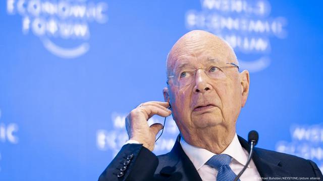 Klaus Schwab sits in front of a lecturn wearing headphones with the right hand raised to his right ear