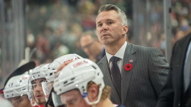 Un entraîneur de hockey derrière le banc lors d'un soir de match regarde quelque chose au loin.