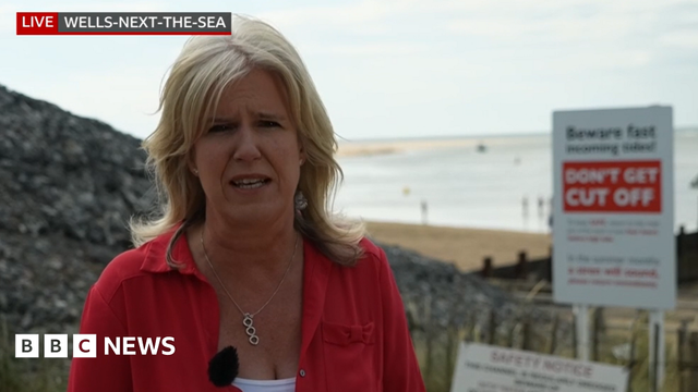 BBC reporter Debbie Tubby, who has shoulder-length blonde hair and is wearing a red jacket over a white top, reports live from a vantage point at the beach. A white and red sign is behind her, warning beach-goers of the dangers of the tide. Sand and water is even further in the background.