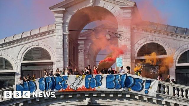 A hand-drawn banner, which says "No Space for Bezos" with a drawing of a rocket ship, is hung along Venice's famous Rialto bridge. A line of people stands behind it, with orange smoke from a flare rising into the sky.