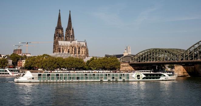 Blick auf den Rhein mit einem Kreuzfahrtschiff darauf, im Hintergrund der Dom und eine Eisenbahnbrücke