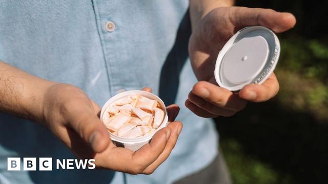 A man shows the camera a small container of nicotine pouches, which look like very small pillows. He is wearing a blue button-down shirt and standing outside.