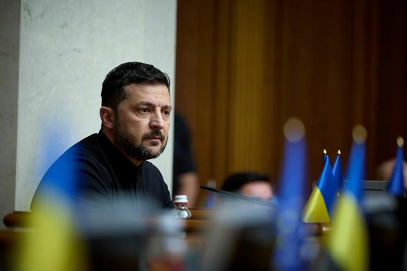 Selenskyj sitzt an einem Pult, vor ihm eine Reihe ukrainischer Flaggen Selenskyj sitzt an einem Pult, vor ihm eine Reihe ukrainischer Flaggen