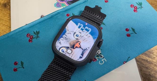 Black Apple Watch Ultra 2 with watchOS 26 loaded on it showing a derpy cat face photo face with liquid glass numbers and a smart stack notification for a camera remote. The watch is on top of a blue notebook with a cherry pattern, that sits on top of a tan notebook with colorful stickers.
