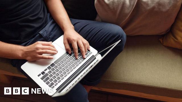 An unrecognisable man wearing a navy t-shirt and dark blue jeans is sitting on a sofa using a laptop.