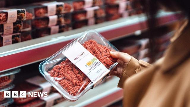 A woman holds a packet of beef mince in a supermarket with rows of mince on shelves in the background