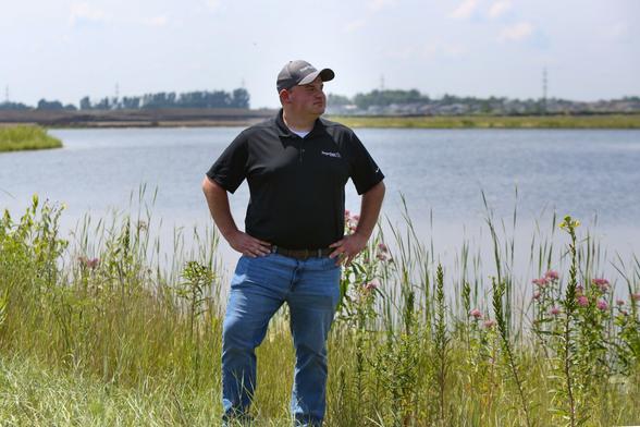 A man in a hat, black polo and jeans stands in front of a body of water.