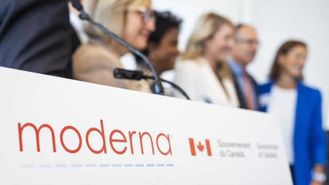 In the foreground: a lecturn with microphones and the logos of Moderna and the Government of Canada, at a press conference on September 19, 2025. Some people are visible in the blurred background.