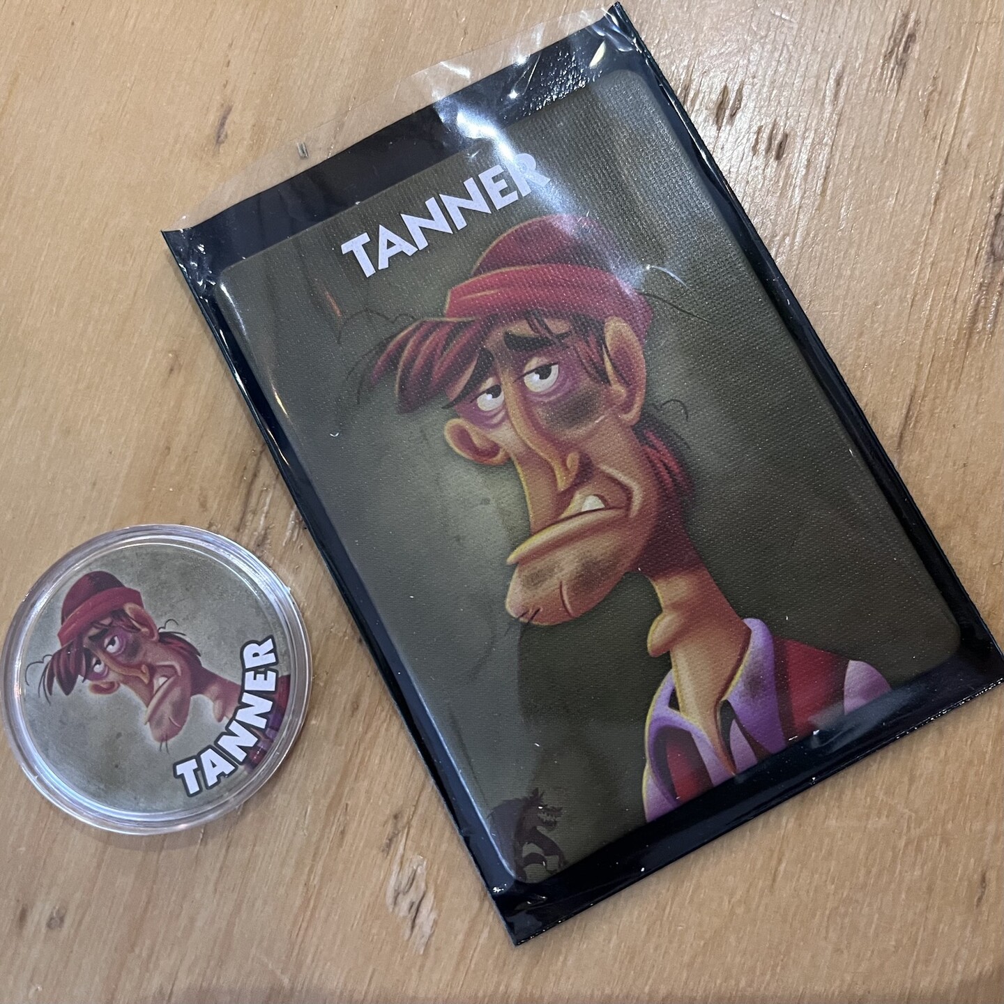 The card and coin of the tanner, a dirty, tired, miserable looking man, from the game One Night Ultimate Werewolf 