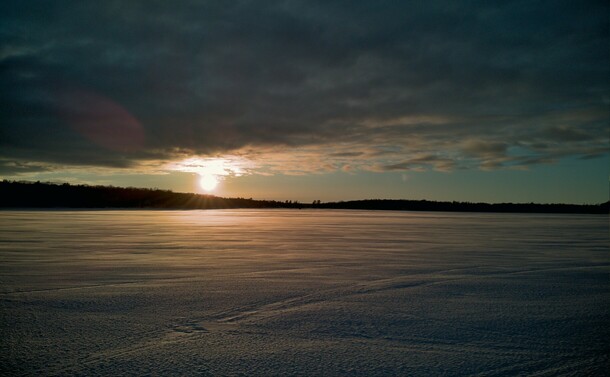 View over snow covered lake. Clouds cover the sky, except for the horizon where the sun is peaking through.