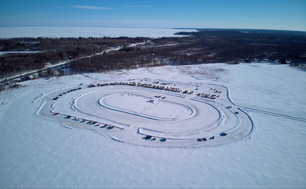 Arial view of a race oval plowed on a frozen lake with about 50-60 cars circled around to watch. Five cars are lined up at start line. 

Larger frozen lake is visible in the background.