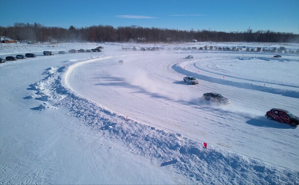 Closer image of a half dozen cars racing on the track, kicking up clouds of snow behind them. Visible around the track are 30-50 cars of spectators watching from inside their cars.