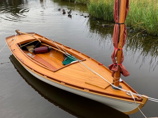 A varnished wooden sailing skiff, with rad tanbark sail. The sail is furled, and the boat is tied to the dock with white lines. Water is calm. Marsh in the background, with ducks.