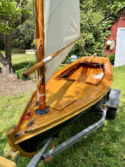 View from above looking down on the deck. Wooden sailing skiff on a trailer in a back yard, in front of an old boat shed. Sunny day, green grass. Cream sail over varnished wood and dark blue hull. 