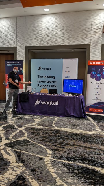 This is a photo of the Wagtail booth at DjangoCon US with a large green banner behind a table with a purple Wagtail tablecloth and stickers and candy on the table.