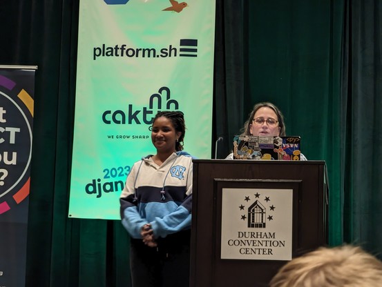 Rachell talking behind the podium as Dawn stands by next to it wearing her university of North Carolina hoodie