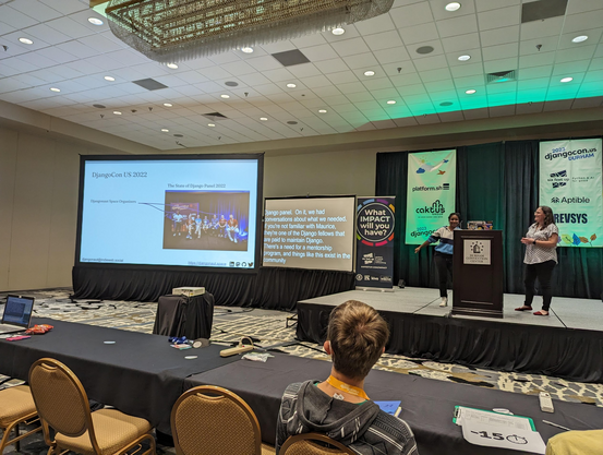 Wide shot of a slide showing the panelists at DjangoCon US 2022 with Rachell and Dawn highlighted, the live captions, and Dawn and Rachell at the podium