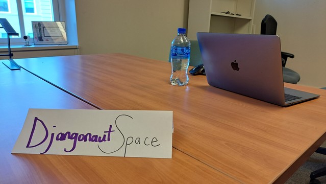 A handwritten sign reading Djangonaut Space. The word Djangonaut is purple. The sign is a paper folded on an office table. There's a computer in the background. 