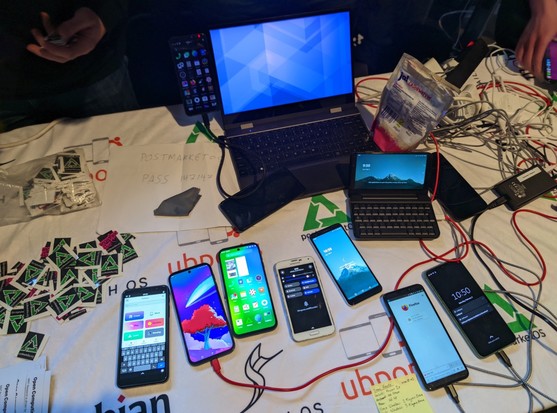 Table with many phones running Linux, a Pinephone with its keyboard case, a Lapdock and many postmarketOS sticksrs