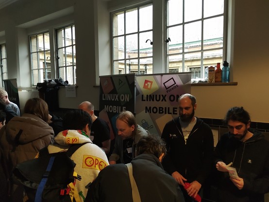 The Linux on Mobile stand with a ton of people around it. It's in a lit room against a wall with high placed windows. Clayton and Pablo are manning the stand for postmarketOS.