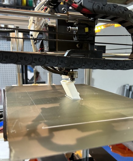 A 3D printer printing a white part. A cable chain is prominent in the foreground.