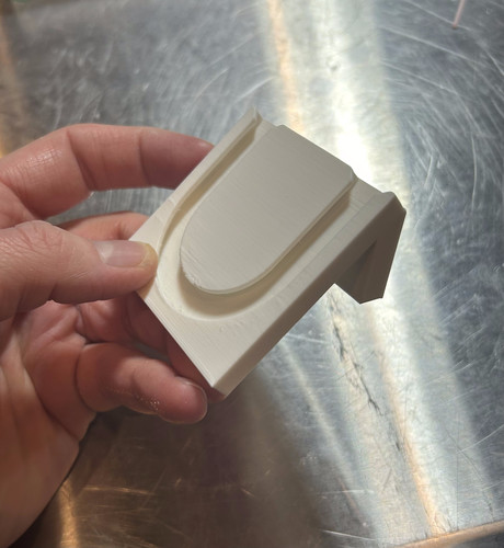 A white 3d printed part, it has an angled front with a u-shaped track in it. It's being held in a hand in front of a shiny metal table.

