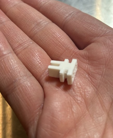 A small white 3d printed clip. It has two prongs in the vertical direction and two in the horizontal direciton at the bottom.
