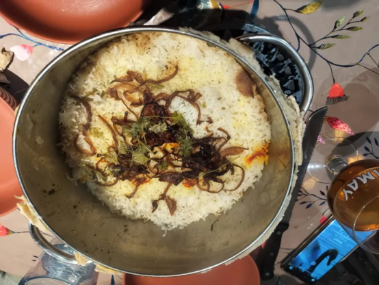 a stainless steel pot in the middle of the table, filled 1/2-way with rice and topped with caramelised onions and herbs. 3 bowls and a glass of beer surround it. 