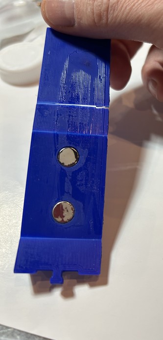 A blue 3d printed object with two magnets held in a hand. 3 folds are visible; on the left the living hinges show discolouring, but are in tact, on the right there is CA glue residue and the hinge is just just gone.