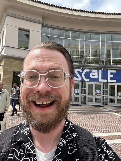 A dork grinning wildly in front of the Pasadena Convention center. There is a blue and white sign that reads SCaLE on the building 