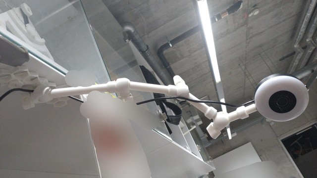 a blurry photo of a lab with a white 3d printed articulated arm with four ball joints, holding a webcam at its end at a complicated angle