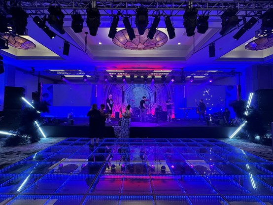 View from the dance floor of the band's stage with some personnel milling, people observing, and blue stage lighting.