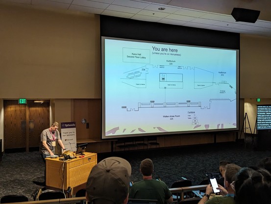 Ben Berry standing on the left of the theater, showing projector screen with layout of the conference venue, titled "You are here"