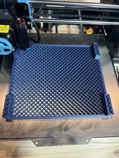 A blue 3d printed object in progress . It has a diamond pattern instead of a top and bottom, there are four towers in the corners. It mostly fill the bed.